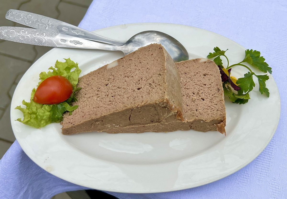 Dwa grube plastry gładkiego pasztetu na białym talerzu z sałatą, pomidorem i natką pietruszki